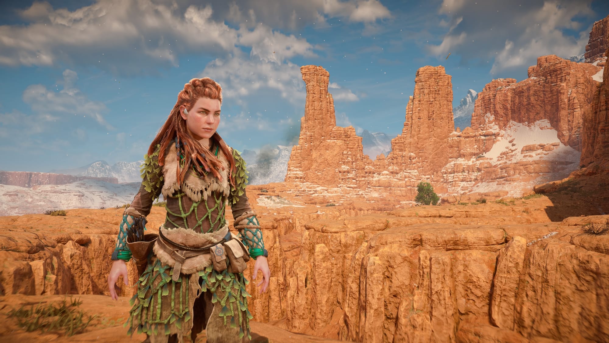 Screenshot from a video game showing a woman with red hair in a woven tribal outfit. Behind her is a visa of red rocks and cloudy sky.
