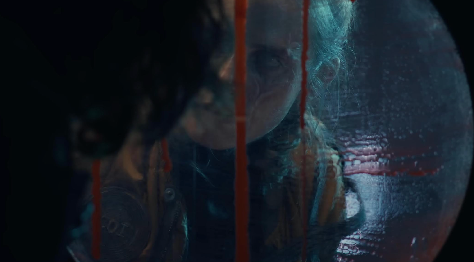 A man peers through a porthole dripping with blood. A woman with a scarred face stares back at him.